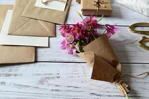 The image shows a rustic wooden table with a small bouquet of delicate pink and purple flowers wrapped in brown craft paper, tied with a piece of twine, creating a charming and natural presentation. photo