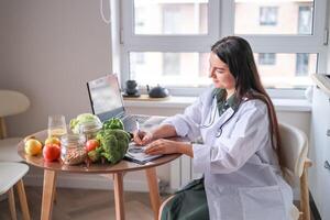 dietitian in a medical coat working with clients online at a table full of fresh vegetables and fruits using a laptop and a tablet. health concept, medical services. photo