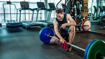 Asian man wiping barbell with red towel in gym after workout, practicing cleanliness and hygiene while holding a cleaner solution in fitness environment photo