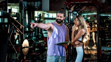 Personal trainer discussing workout strategy with female client at the gym, pointing towards equipment while planning fitness goals with clipboard in hand photo