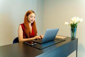 Asian woman working remotely from home on laptop while sitting at a modern desk with flower vase, smiling and focused in a minimalist workspace setup. photo