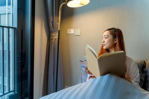 Young woman reading a book while sitting comfortably in bed near window with soft lighting, creating a cozy and peaceful morning atmosphere in a modern room photo