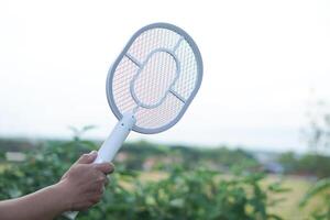 White electric mosquito swatter held firmly in hand, ready to zap away annoying insects in outdoor background. Modern, innovative device designed to eliminate mosquitoes and flying pests effectively. photo