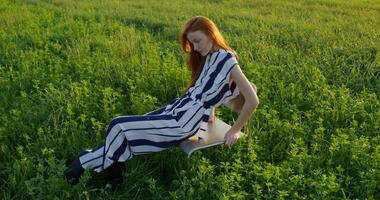 A red-haired woman poses looking at the camera while sitting on a chair in the middle of a field, she smiles. A sunny summer day photo