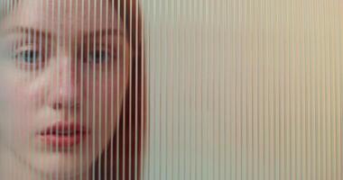A red-haired woman with freckles looks at the camera with a serious look standing behind striped plexiglass in the studio photo