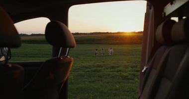 Three young friends take from the side of the field to the car at sunset. The view from the car window. Summer evening photo