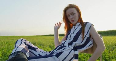 A red-haired woman poses looking at the camera with a calm look while sitting on a chair in the middle of a field. A sunny summer day photo