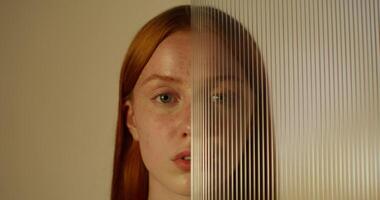 A red-haired woman with freckles looks at the camera with a serious look standing on a studio background. Half of her face is covered with plexiglass photo