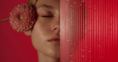 The woman looks at the camera with a serious look and touches her face with a flower. Half of her face is behind a glass with water running down it photo