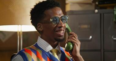 A man is talking on a wired phone while sitting in a retro office. He's smiling. photo