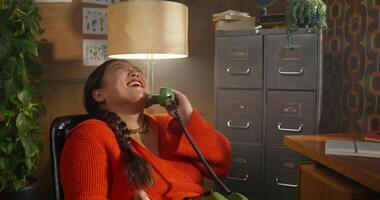 A woman is talking on a wired phone while sitting in a retro room. She's laughing. photo