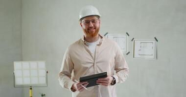 A man in a construction helmet and with a tablet in his hands is looking at the camera standing in a room without decoration photo