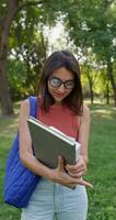 A young woman with glasses is smiling at the camera while standing in a city park. She holds a notebook and a book in her hands. Summer day photo