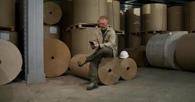 A man uses a mobile phone at a paper mill. He's sitting on a roll of paper in a warehouse photo