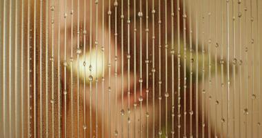 A woman with eye patches looks at the camera with a serious look standing behind a glass with water running down it. She touches her face with a flower photo