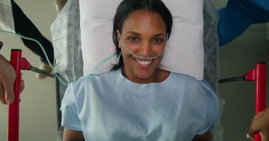 A woman with an oxygen tube in her nose smiles at the camera while lying on a medical gurney. She is being driven down the hospital corridor photo