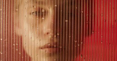 A woman looks at the camera with a serious look standing behind a glass with water running down about it in the studio. Close-up of her face photo