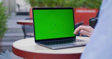 A man uses a laptop with green screen sitting at a table on a cafe terrace. Close-up of his hands, an unrecognizable persona. Copy space photo