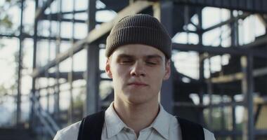 A young man of European appearance in a knitted hat with a backpack on his back looks straight into the camera with a serious look, portrait photo