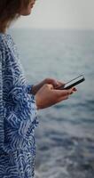 A woman is typing on a mobile phone while standing on the rocks on the seashore. No face, vertical . The woman is blurry. 4k , red komodo photo