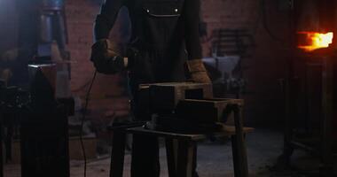 A blacksmith of African American appearance in a leather apron and gloves cuts off a piece of metal workpiece using an angle grinder. close-up, no face. 4k, red komodo photo