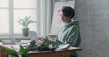 A young serious woman is typing on a laptop while sitting at a desktop in an art workshop, with drawings and paints laid out on it photo
