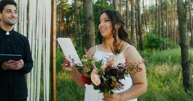 The bride laughs while reading the oath standing opposite the groom in front of the altar with the priest in the forest photo