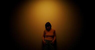 A woman in a wig is leaning against the wall posing in the orange light of a lamp in the studio photo