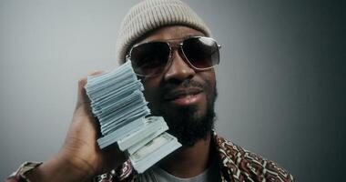 A bearded man in a hat and sunglasses, with a wad of money in his hand, smiling at the camera standing on a gray background in the studio. Dollar bills are falling on top of him photo