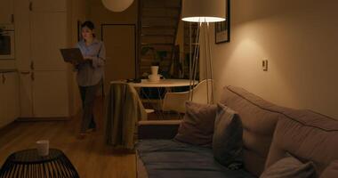 A woman with an open laptop in her hands sits down on the sofa and starts typing. Dimmed evening light in the apartment photo