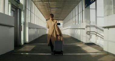 A woman in formal clothes and a coat uses a mobile phone walking along the corridor of the railway station, rear view photo