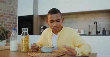 The boy has breakfast at home, he eats cereal with milk sitting at the table and looking at the tablet photo