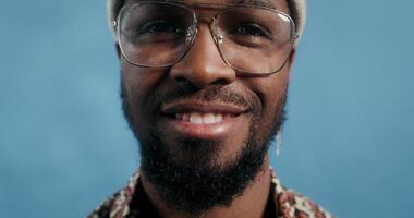 A bearded man with glasses and a hat, with an earring in his ear, smiling at the camera standing on a blue background in the studio, close-up of his face photo