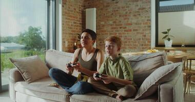Mom and son of European appearance playing games using joysticks sitting on the couch in a stylish home interior. They rejoice in victory photo