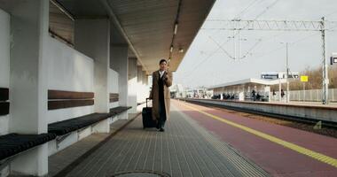 A woman in formal clothes and a coat uses a mobile phone walking along the station platform with a suitcase in her hand photo