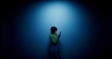A woman in a wig uses a mobile phone, leaning against the wall in the blue backlight in the studio photo
