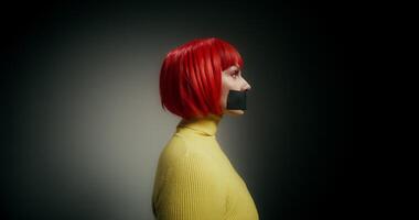 A woman in an orange wig with her mouth taped shut turns her head and looks at the camera with a frightened look photo