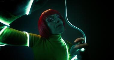 A woman in an orange wig and glasses, wrapped in an LED strip, dances looking at the camera on a studio background photo