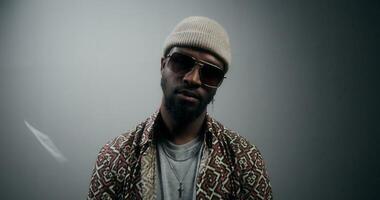 A bearded man in sunglasses and a hat, with jewelry, looks at the camera with an independent look, standing on a gray background in the studio. Dollar bills are raining down on him photo