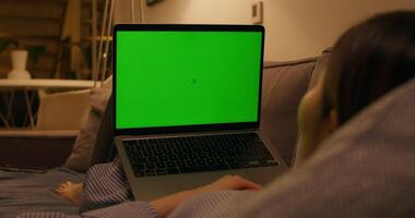 A woman uses a laptop with green screen lying on the couch in an apartment with dim lights, close-up of the screen, an unrecognizable person photo