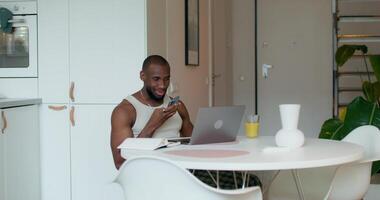 A man is smiling while recording a voice message on a mobile phone sitting at a table in the kitchen. An open laptop and a notebook in front of him photo
