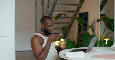 A man is recording a voice message on a mobile phone sitting at a table in the kitchen. An open laptop and a notebook in front of him photo