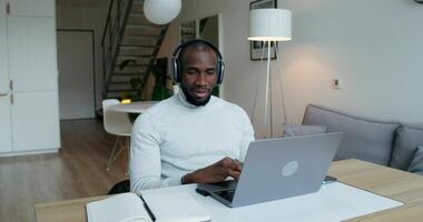 A man with headphones waves hello starting a chat on a laptop sitting at a table at home. He writes in a notebook. Remote work photo