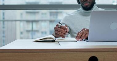 A man with headphones is writing in a notebook sitting at a table in front of an open laptop near the office window photo