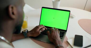 A man uses a laptop with a green screen sitting at a table at home, rear view, unrecognizable person. Daylight photo