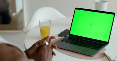 A man is talking via chat on a laptop with a green screen sitting at a table at home. Rear view, unrecognizable person photo