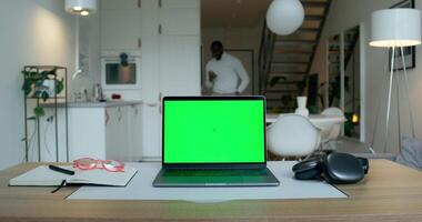 A laptop with a green screen is on the desktop at home. A man uses a mobile phone standing in the kitchen in the background photo