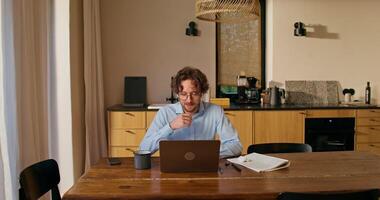 A man with glasses and earbuds is talking on a chat on a laptop and drinking coffee sitting at a table in the kitchen at home photo