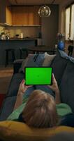 A woman is holding a tablet with green screen lying on the couch at home, an unrecognizable person photo