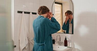 A man in a bathrobe applies moisturizing patches to his eyelids while standing in front of a bathroom mirror photo